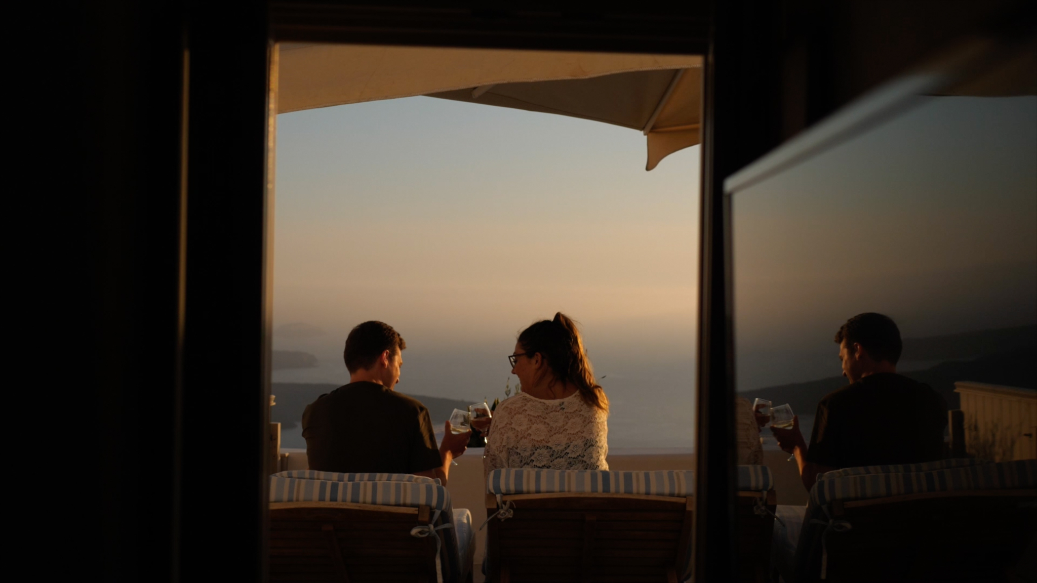 Sara and Gareth cheers to their wedding day while sitting on their balcony in Santorini. Their reflection can be seen in the window, and the view over the sea can be seen in the distance.