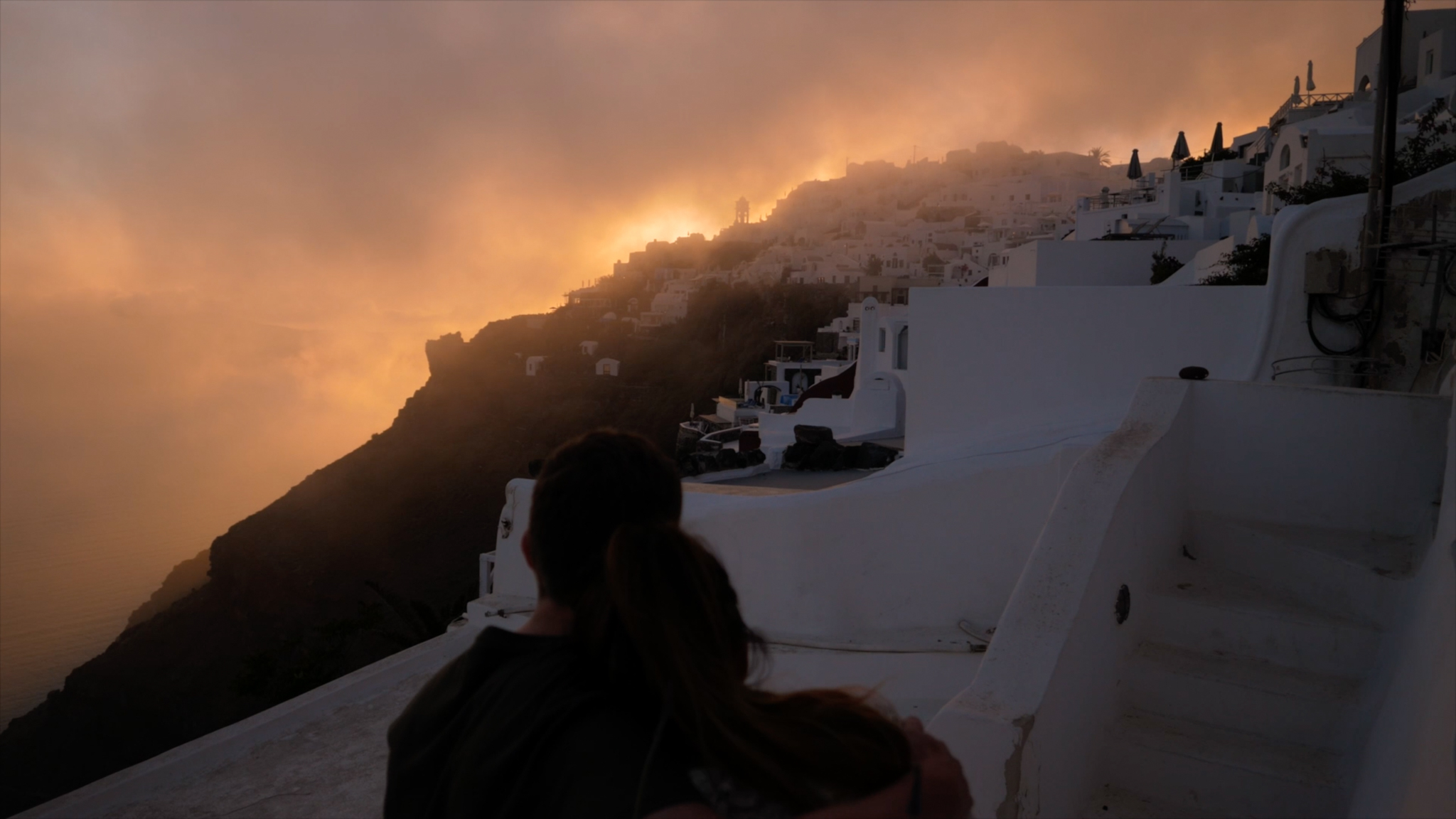 A shot of the houses on Santorini hills with the couple sitting on the steps to their villa watching the sunset as it falls behind the hills, disappearing into the low clouds.