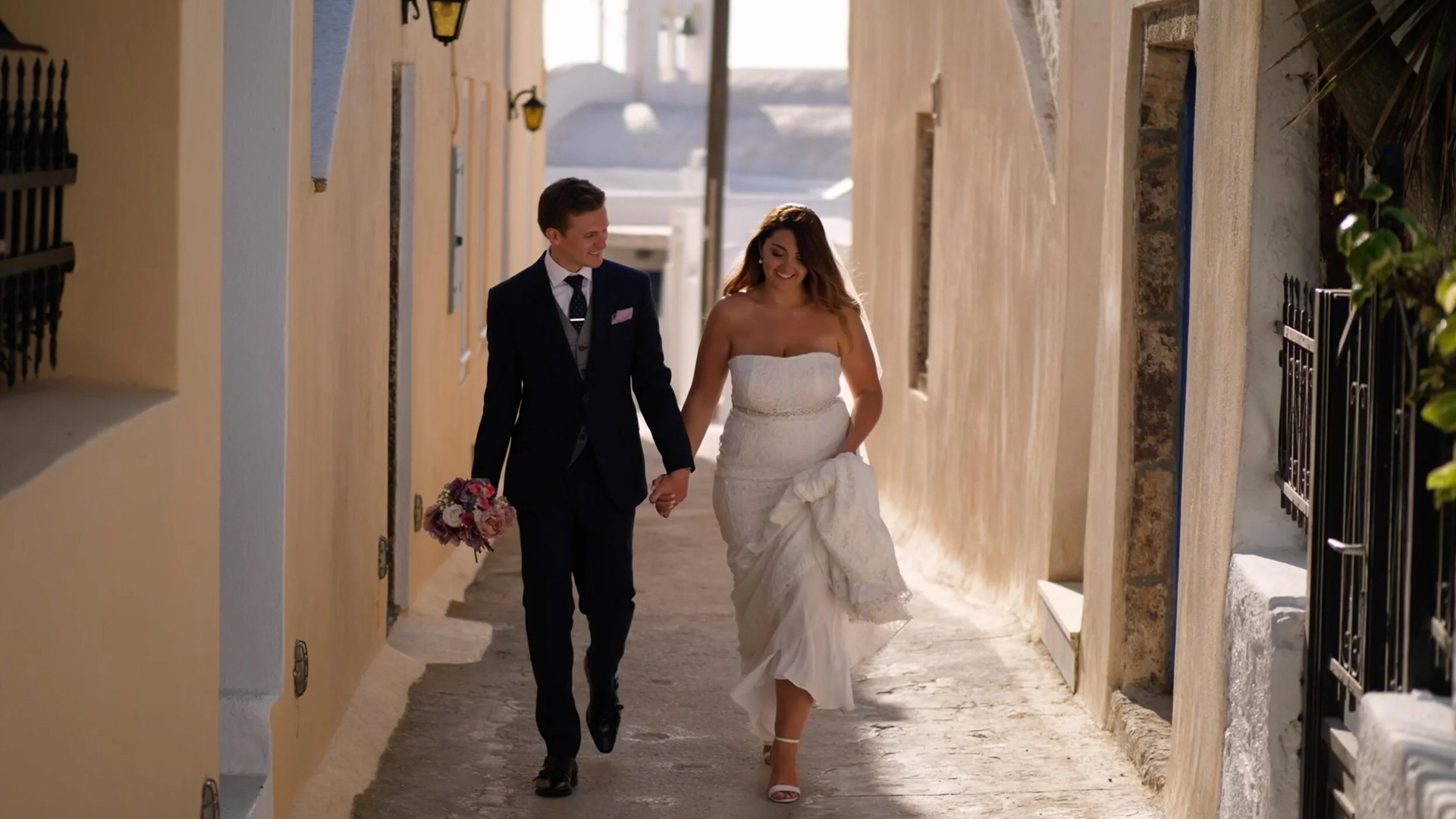 The Bride and Groom take a stroll through the streets of Santorini, Greece. The sunlight shines behind them, creating a halo of sorts around the bride, as she holds her husband's hand. Out-of-focus foliage can be seen on the right of the frame, as the buildings disappear behind them with leading lines pulling you into the frame.