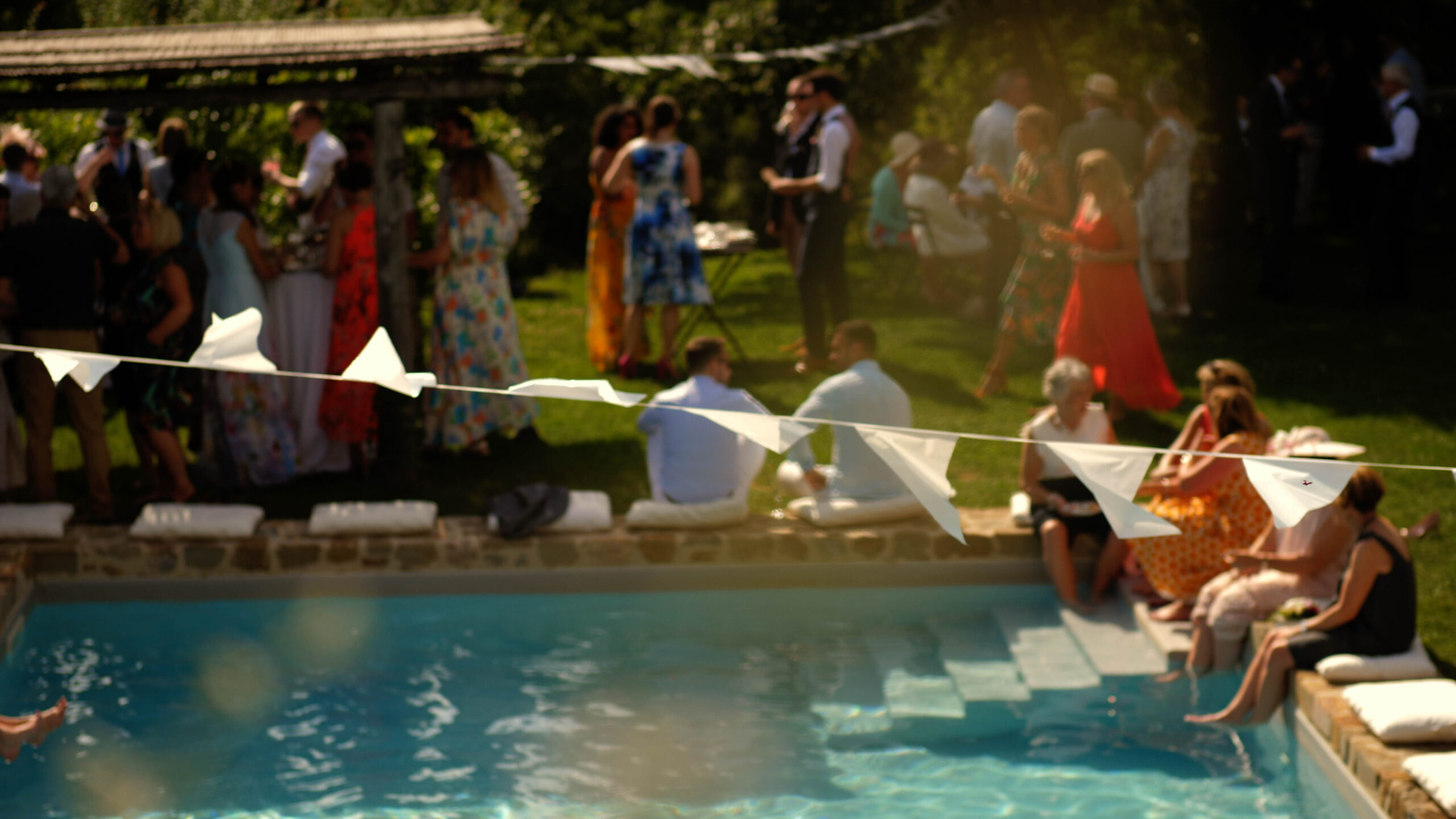 Beth and Steve's pool party at Villa Podernovo, Tuscany, Italy. Image shows the swimming pool and guests celebrating their wedding day. The focus is on the banner, and the guests are out of focus in the background.