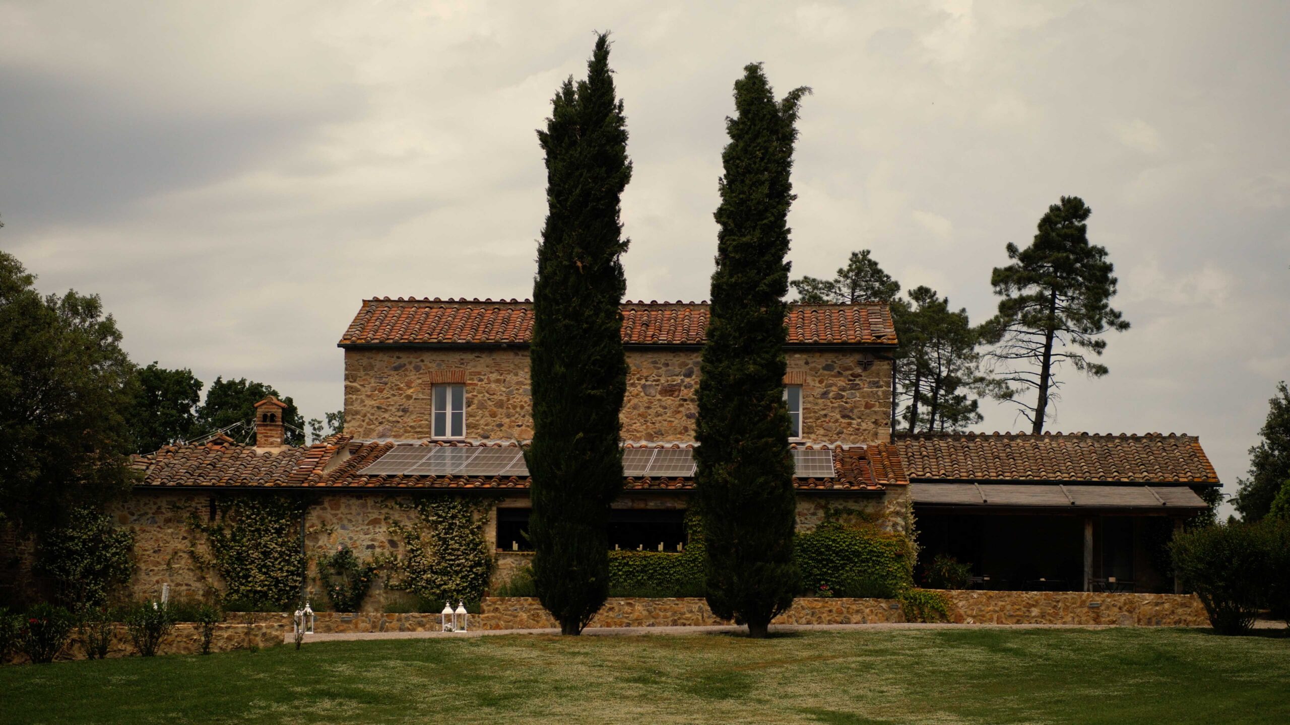 A shot of Villa Podernovo, Tuscany, Italy. Two trees stand tall in front of the villa, which is surrounded by beautiful trees.