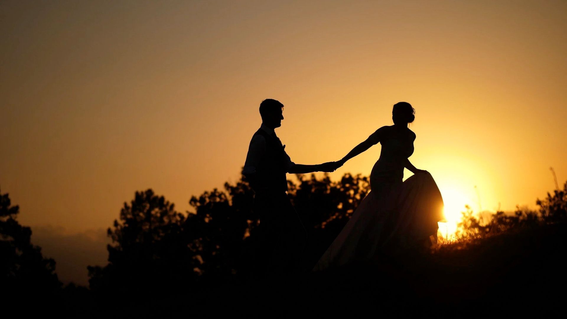 Silhouette of the bride and groom walking on the hills of Villa Podernovo in Tuscany, Italy. The sunset is behind them, as the bride's dress moves through the sun, creating an orange glow around her dress. The couple are walking hand in hand, silhouetted against the orange sky.