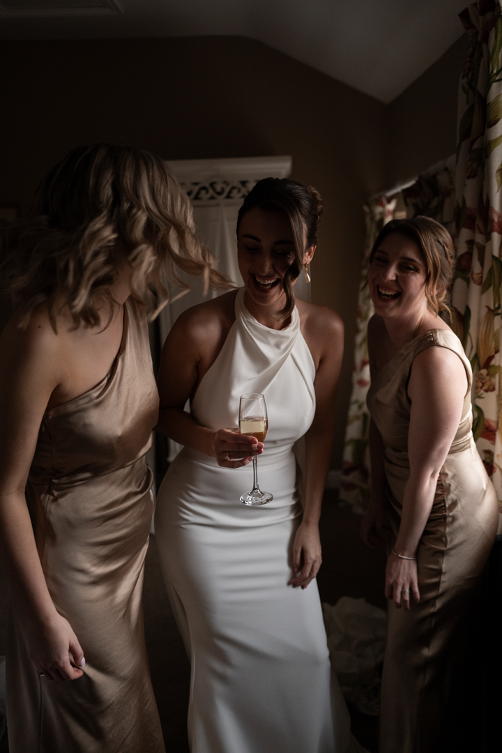 Bride takes a moment to have a glass of champagne with her bridesmaids during preparations. The girls are laughing, and the light is pouring through from the window onto the bride and her dress.
