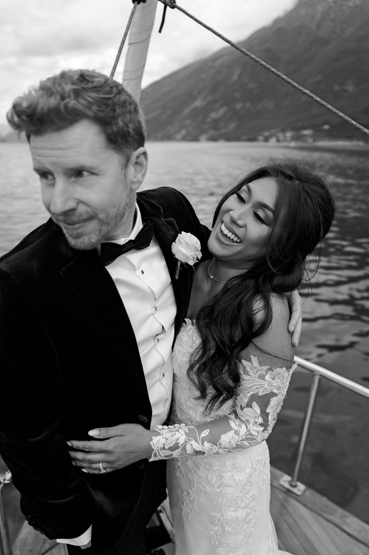 The bride and groom stand at the front of a boat while sailing on Lake Garda in Italy. After getting married at Malcesine Castle, the couple are happy with the bride laughing, and the groom admiring the views. They have their arms wrapped around one another, and the shot is in black and white, showing a part of the boat, the hills and the lake itself.
