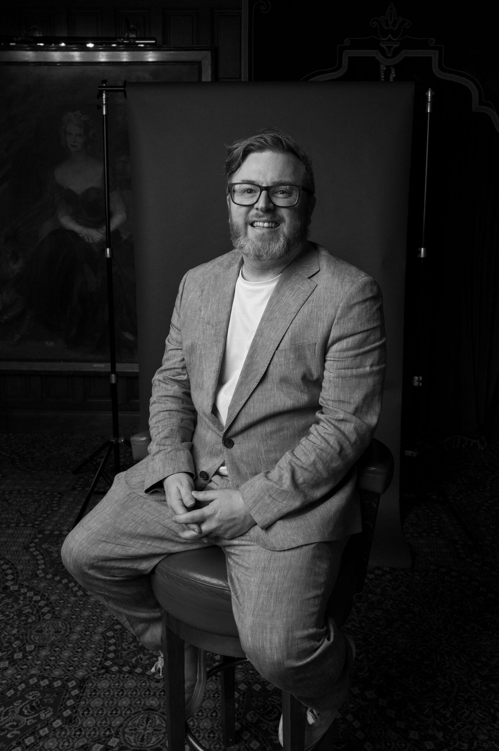 Craig is captured while sitting on a stool in front of a professional backdrop. He is having his photograph taken by Philip White, as they capture a wedding at Ashford Castle in Ireland. Craig has his fingers held together with both hands, while his feet swing back around the stool. Craig has a beaming smile, looking directly into the lens of the camera.