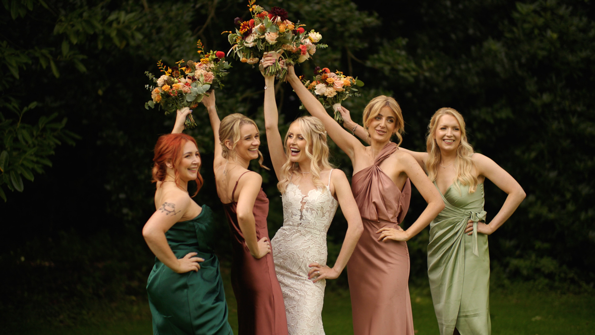 The bride and her bridesmaids hold their bouquets aloft as they pose for a group photograph in front of the trees on their wedding day. The bride and her bridesmaids are all smiling or laughing and the bouquets make a nice triangle hanging above their heads.