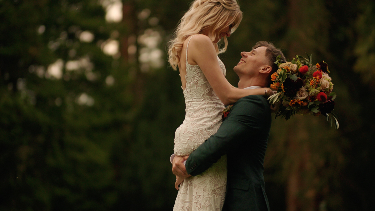 A groom wearing a green suit lifts his bride in front of the forest, as they each admire one another. The bride's bouquet hangs behind the groom's back, as they lean in for a kiss.